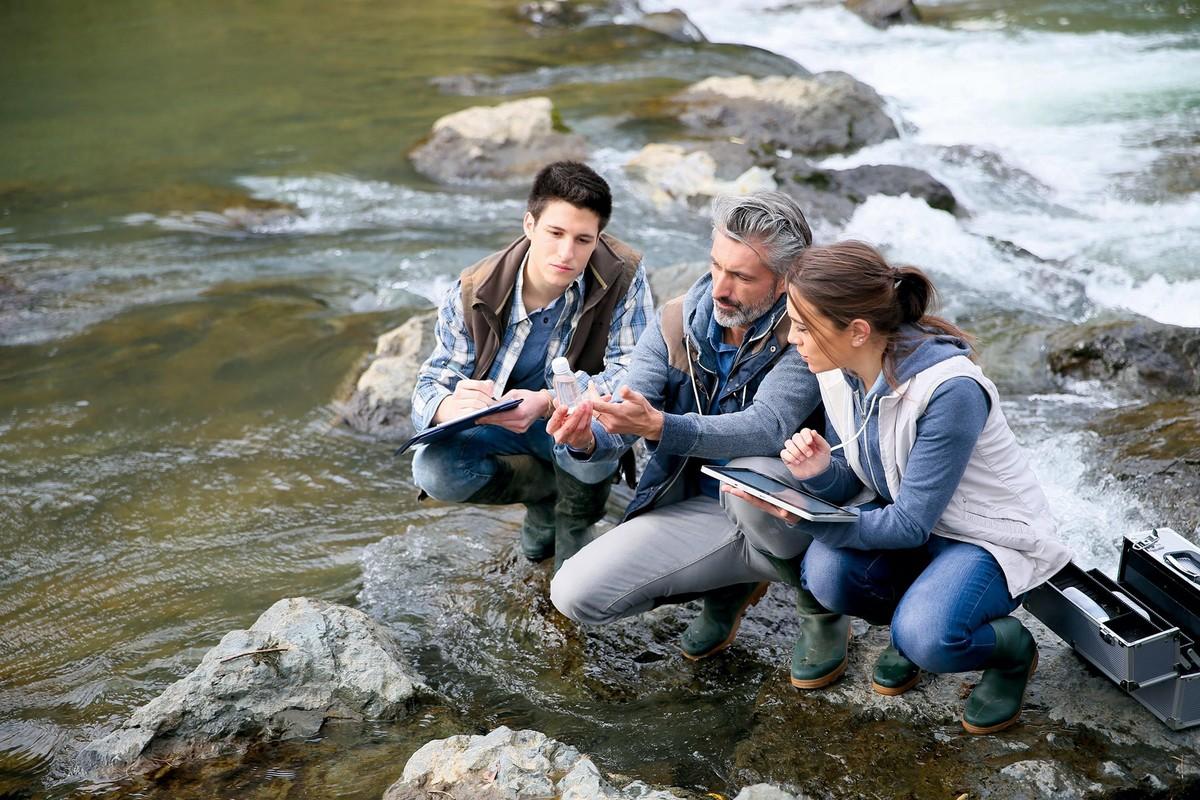 Trois personnes au bord d'une rivière entrain d'effectuer le suivi de la biodiversité dans une rivière.